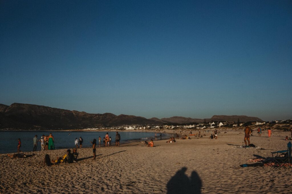 Menschen stehen verstreut an einem Sandstrand bei Sonnenuntergang, mit Bergen im Hintergrund, Häusern in der Ferne und einem klaren, blauen Himmel über uns. Im Vordergrund sind die Schatten von zwei Figuren zu sehen - die perfekte Inspiration für jeden Fotograf Köln oder Werbefotograf.