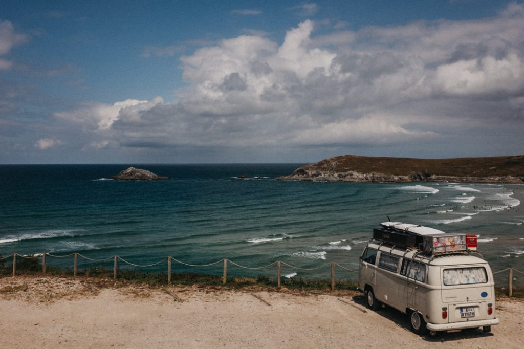 Ein altes weißes Wohnmobil mit Gepäck auf dem Dach steht an einem Aussichtspunkt an der Küste, ideal für einen Familien- oder Kinderfotografen, der blaue Wellen, eine kleine Insel und sanfte Hügel unter einem teilweise bewölkten Himmel festhält.