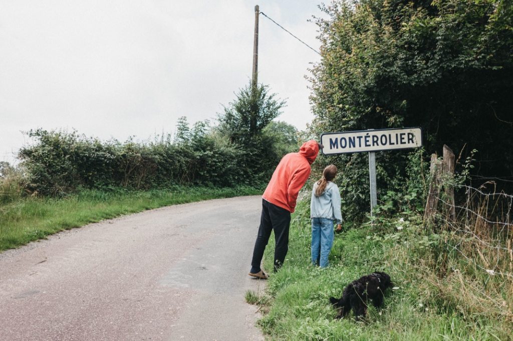 Eine Person in einer roten Jacke und ein Kind betrachten ein Straßenschild von Montérolier an einem ländlichen Straßenrand, mit einem schwarzen Hund in der Nähe - eingefangen von einem Kinderfotografen, der an Schnappschüsse aus einem Fotostudio Köln erinnert. Der Hintergrund ist begrünt.