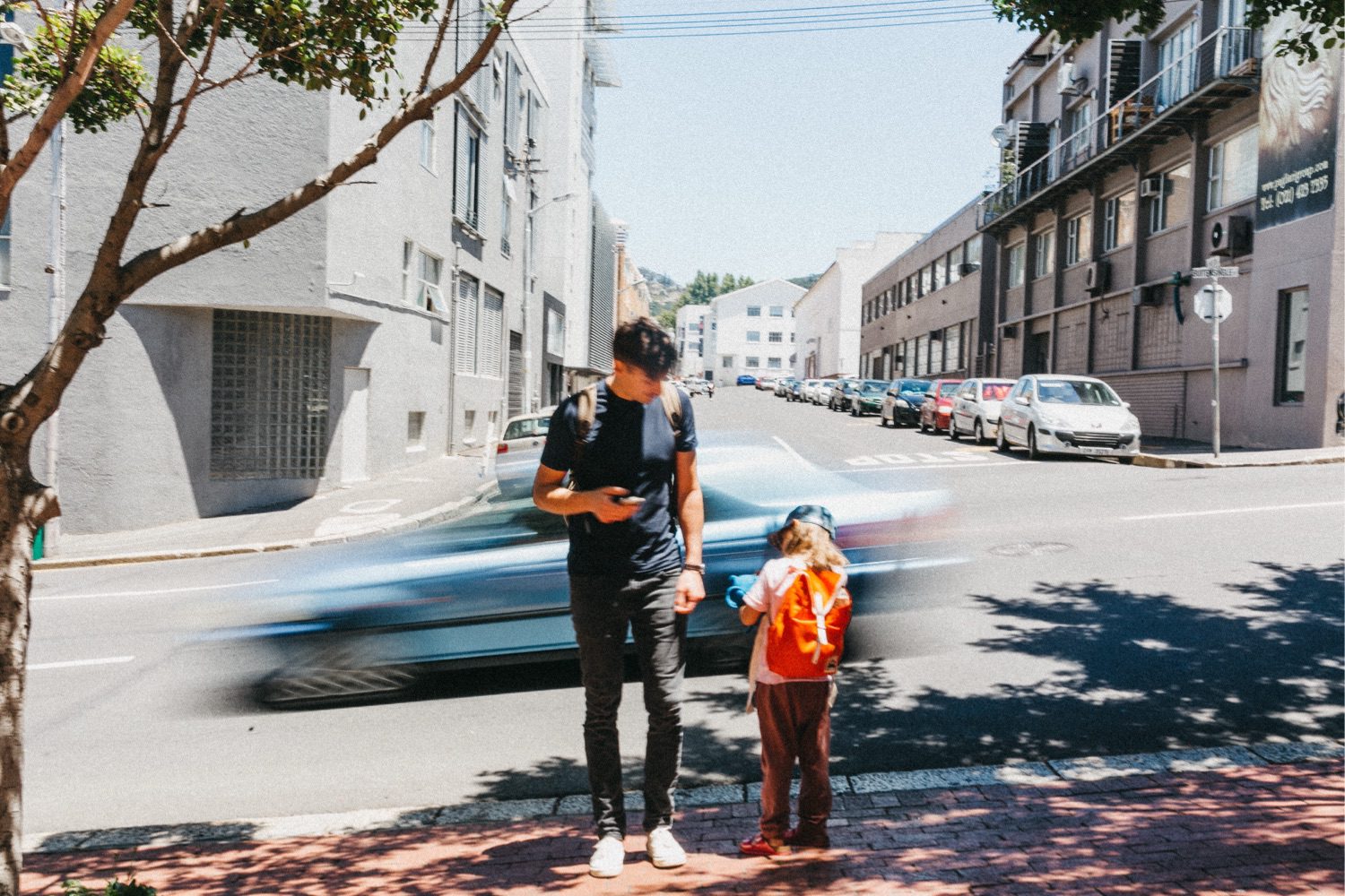 Ein Mann und ein Kind mit einem leuchtend orangefarbenen Rucksack stehen auf einem Bürgersteig. Aufgenommen von einem talentierten Kinderfotografen, prüft der Mann sein Handy, während ein blaues Auto auf einer sonnigen Straße mit geparkten Autos und Gebäuden unscharf vorbeifährt.