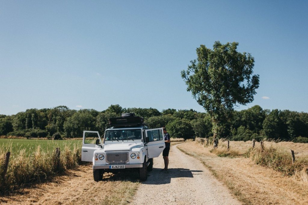 Ein weißer Geländewagen mit offenen Türen parkt auf einem ländlichen Feldweg neben einem Feld und einem großen Baum, und eine Person - möglicherweise ein Werbefotograf - steht in der Nähe unter dem klaren blauen Himmel.
