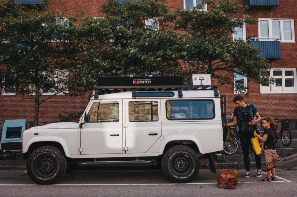 Ein Mann und ein junges Mädchen, aufgenommen von Kids Fotograf, packen ihre Ausrüstung in einen weißen Geländewagen mit der Aufschrift "KAMPER", der auf einer Straße vor einem Backsteinwohnhaus geparkt ist. Eine braune Tasche liegt neben ihnen auf dem Boden.