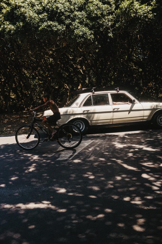 Eine Person fährt mit dem Fahrrad auf einer sonnenbeschienenen Straße an einem geparkten cremefarbenen Oldtimer vorbei, während hohe, belaubte Bäume im Hintergrund Schatten werfen - ein ideales Motiv für jeden Familien- oder Werbefotografen, der authentische Momente sucht.