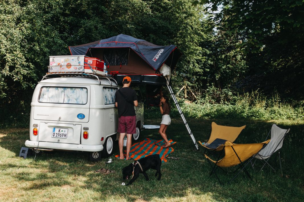 Ein weißes Wohnmobil mit einem Dachzelt und Gepäck ist auf einer Wiese geparkt. Zwei Menschen und ein schwarzer Hund sind in der Nähe. Gemütliche gelbe Stühle und eine gestreifte Decke stehen unter grünen Bäumen - perfekt für ein entspanntes Kinderfotograf- oder Fotografin-Shooting im Freien.