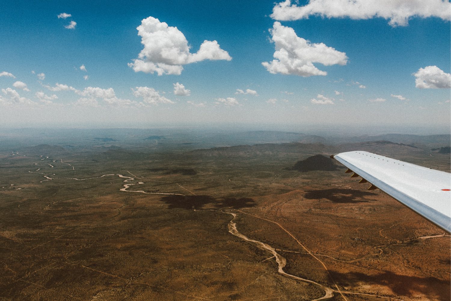 Luftaufnahme einer trockenen Landschaft mit gewundenen Flüssen und zerklüftetem Gelände, gesehen aus einem Flugzeugfenster mit einem Teil der Tragfläche unter einem blauen Himmel mit vereinzelten Wolken - aufgenommen von einem Familienfotografen aus dem Fotostudio Köln.
