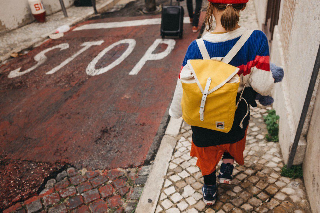 Eine Person mit einem gelben Rucksack geht auf einer Kopfsteinpflasterstraße neben einem aufgemalten STOP-Schild. Sie trägt einen bunten Pullover und einen roten Hut und zieht einen kleinen Koffer vor sich her. Die urbane Szene spiegelt den offenen Stil eines Familienfotografen wider.