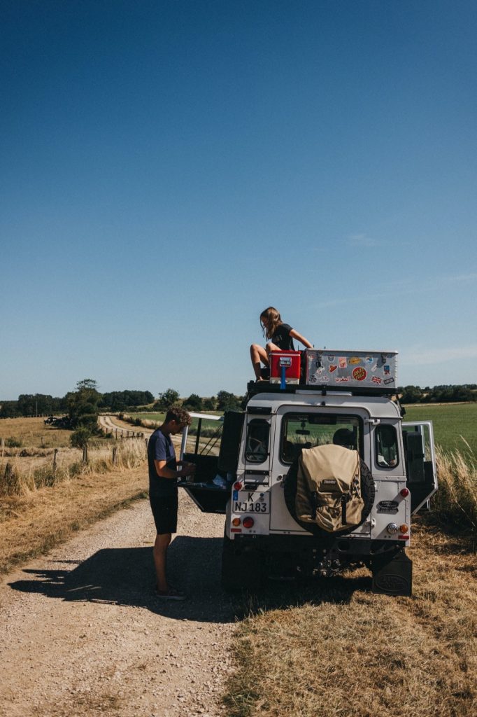Eine Fotografin steht neben einem weißen Geländewagen, der auf einem ländlichen Feldweg geparkt ist, während eine andere Person auf dem Dach unter dem klaren blauen Himmel sitzt. Im Hintergrund erstrecken sich Felder und Bäume, die Momente wie diese von Fotograf Köln einfangen.