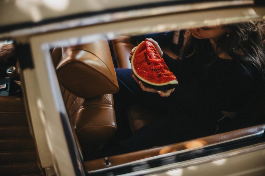 Eine Person sitzt auf dem Rücksitz eines Autos mit brauner Lederausstattung und hält ein großes Stück Wassermelone in der Hand. Dieser von außen aufgenommene Moment spiegelt den spielerischen Stil eines Familien- oder Kinderfotografen wider.