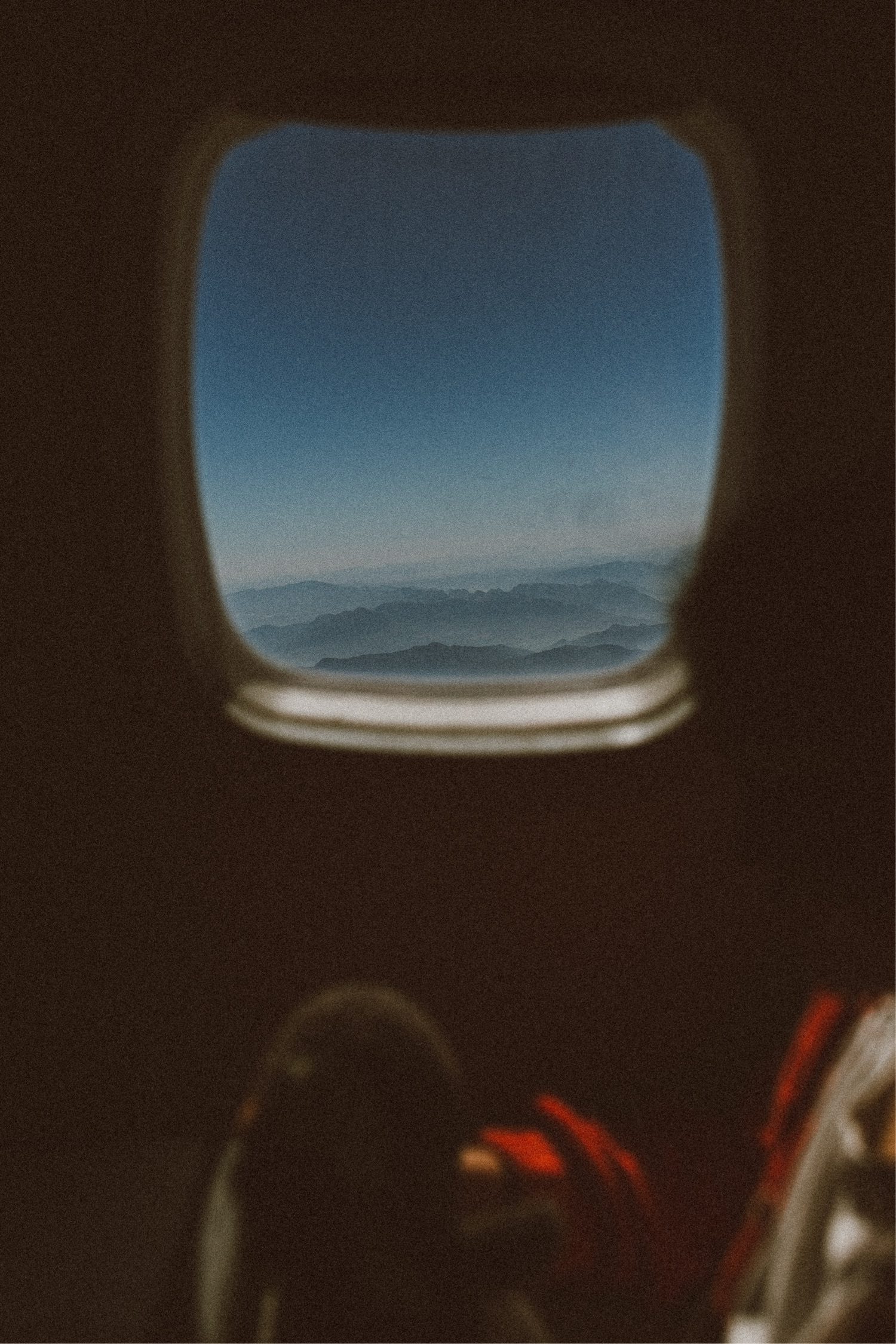 Blick aus dem Fenster eines Flugzeugs mit dunstigen blauen Bergen unter klarem Himmel, aufgenommen im Stil eines Werbefotografen, mit der unscharfen Silhouette eines sitzenden Passagiers im Vordergrund.