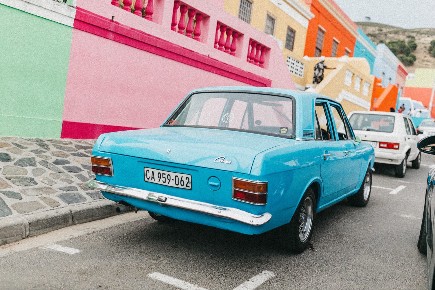 Ein leuchtend blauer Oldtimer parkt auf einer Straße, die von bunten Häusern in Rosa-, Orange- und Blautönen gesäumt ist - eingefangen von einem Werbefotografen -, während andere Fahrzeuge ebenfalls den Bordstein säumen.