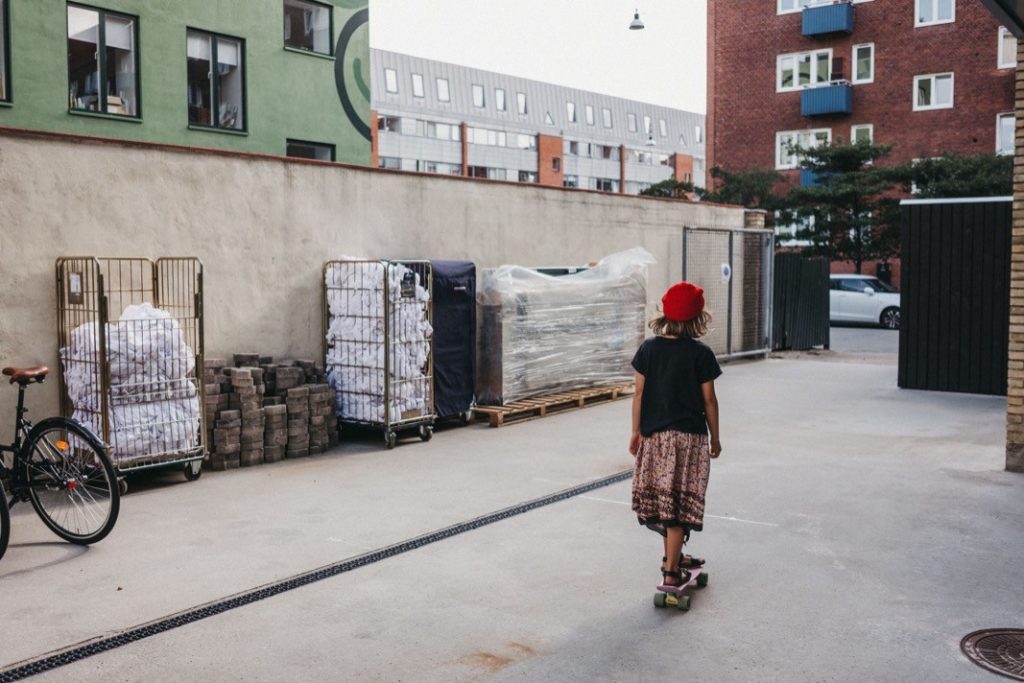 Ein kleines Kind mit roter Mütze und gemustertem Rock fährt auf einem Skateboard durch eine urbane Gasse, neben Stapeln von verpackten Waren und einem geparkten Fahrrad, mit Wohnhäusern im Hintergrund - aufgenommen von einem talentierten Kinderfotografen aus dem Fotostudio Köln.