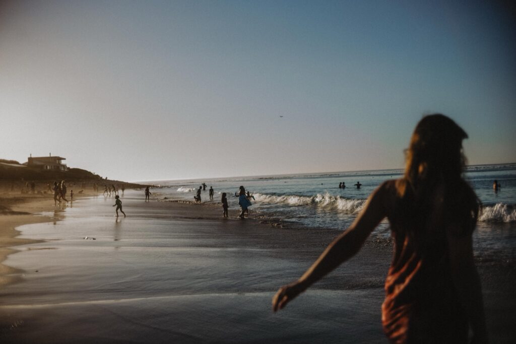 Menschen genießen einen sonnigen Tag am Strand; einige schwimmen im Meer, während andere auf dem nassen Sand spazieren gehen oder stehen. Die Aufnahme einer talentierten Fotografin zeigt eine verschwommene Figur im Vordergrund, während in der Ferne weitere Menschen und ein Haus zu sehen sind.