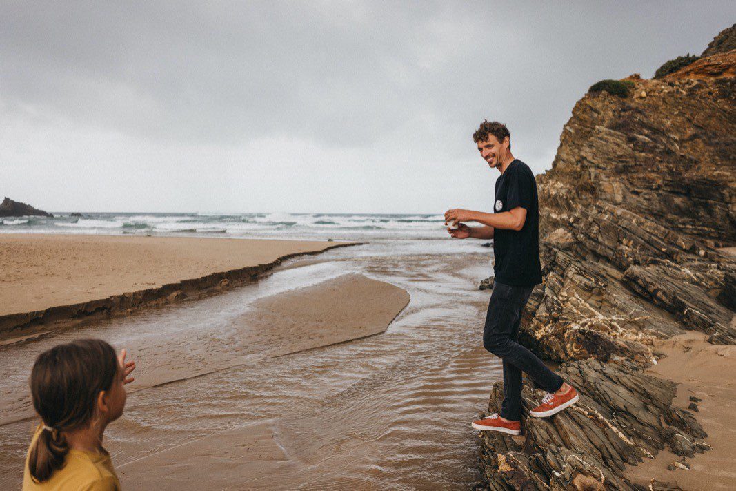 Ein Mann in schwarzem Hemd und roten Schuhen steht auf Felsen an einem Bach, der auf den Strand trifft, und lächelt ein Kind in einem gelben Hemd an - wunderschön eingefangen von einem Familienfotografen mit dem Meer und dem bewölkten Himmel im Hintergrund.