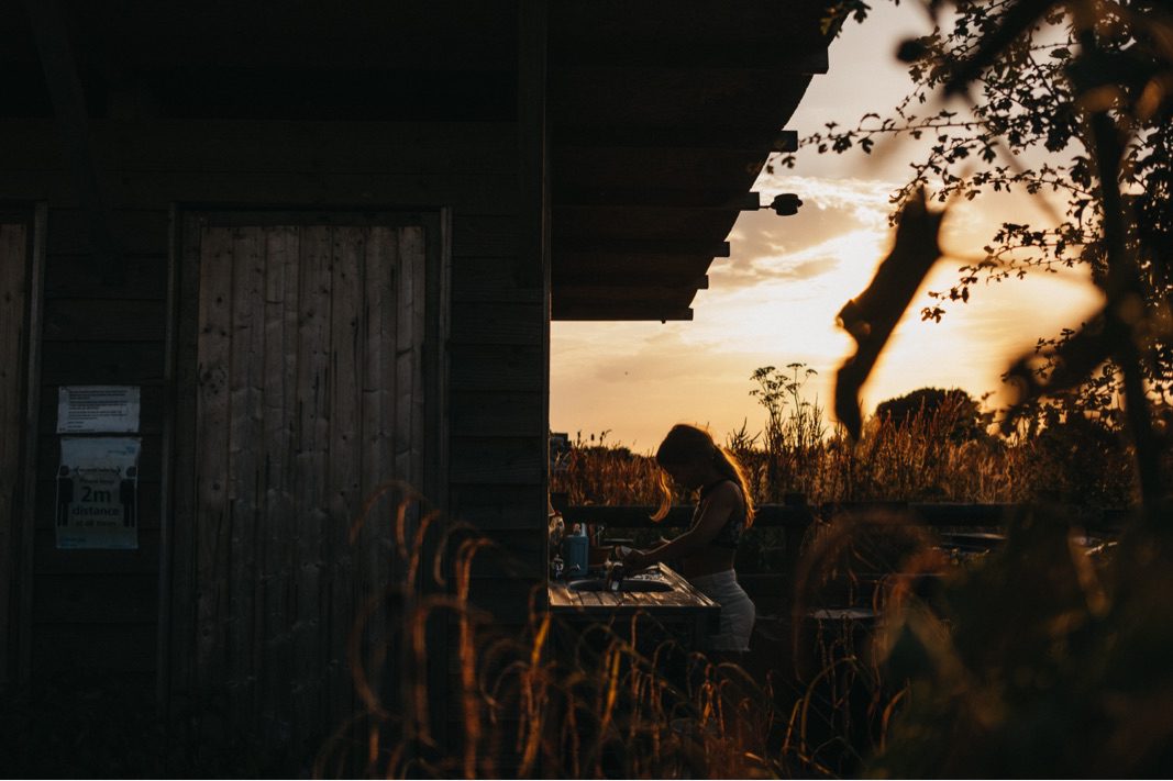 Eine Frau bereitet an einer Theke im Freien neben einem Holzgebäude bei Sonnenuntergang Essen zu, festgehalten von einem talentierten Werbefotografen. Hohes Gras und Pflanzen füllen den Vordergrund, der in das warme, goldene Licht des Himmels getaucht ist.