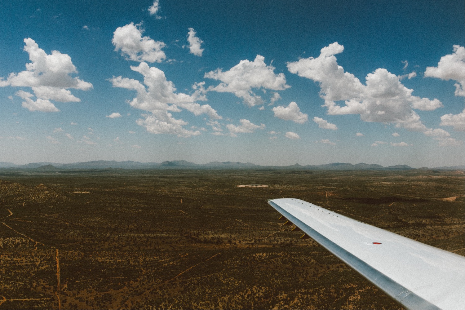 Luftaufnahme einer weiten, grünen Landschaft mit spärlicher Vegetation, gesehen aus dem Fenster eines Flugzeugs. Aufgenommen von einem talentierten Familienfotografen, ist rechts die Tragfläche des Flugzeugs unter einem blauen Himmel mit vereinzelten weißen Wolken zu sehen.