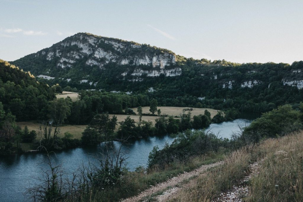 Ein sich schlängelnder Fluss fließt durch ein üppig grünes Tal, gesäumt von dichten Bäumen und grasbewachsenen Feldern. Im Hintergrund erhebt sich ein felsiger, bewaldeter Berg - ein ideales Motiv für einen Familienfotograf oder eine Fotografin aus Köln, um es unter dem klaren Himmel festzuhalten.