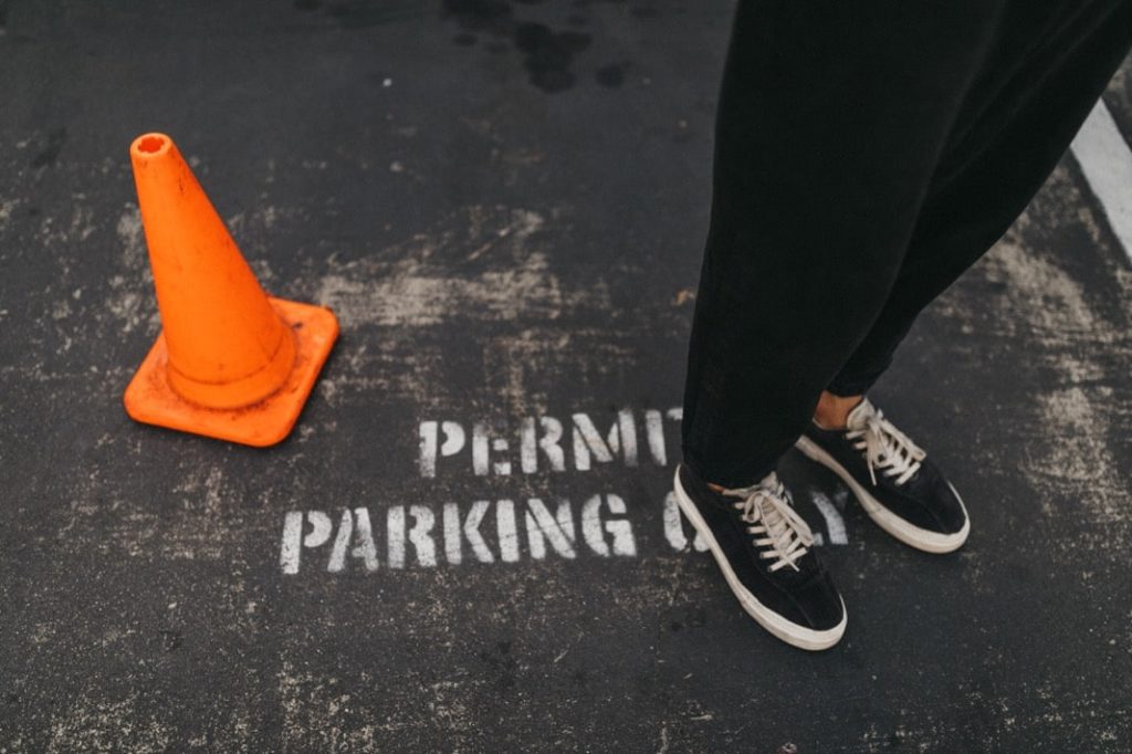 Ein Werbefotograf fängt eine Person in schwarzer Hose und Turnschuhen ein, die neben einem orangefarbenen Verkehrskegel auf dem Asphalt steht, auf dem in weißer Farbe PERMIT PARKING ONLY steht.