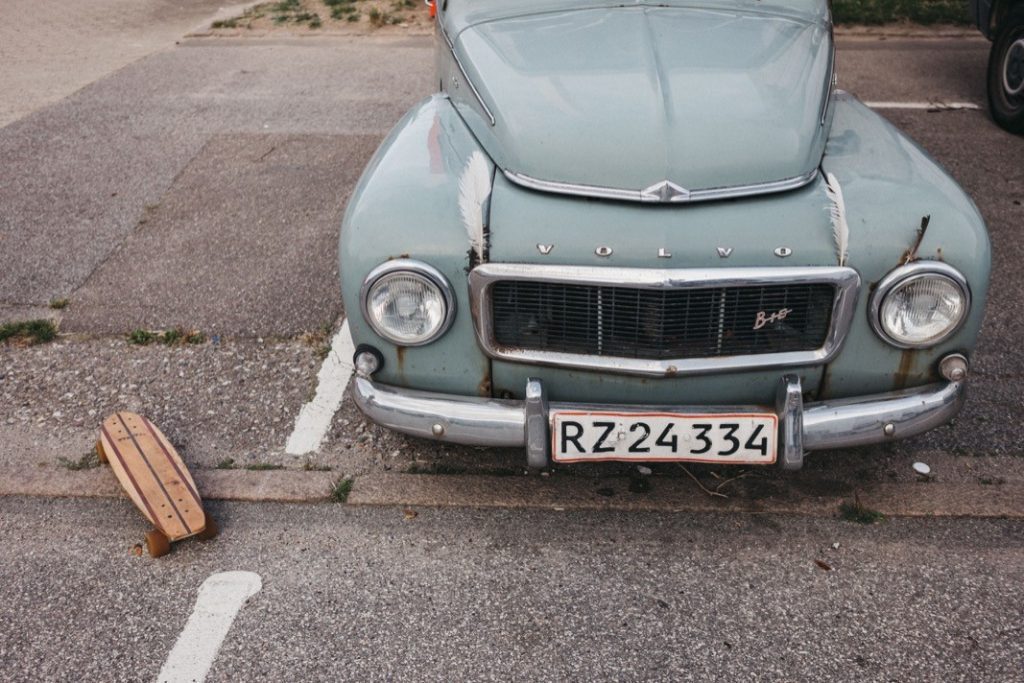 Ein hellblauer Volvo-Oldtimer mit sichtbaren Rostflecken ist auf einem Parkplatz neben einem hölzernen Skateboard geparkt. Das von einem Werbefotografen aufgenommene vordere Nummernschild lautet RZ 24334.