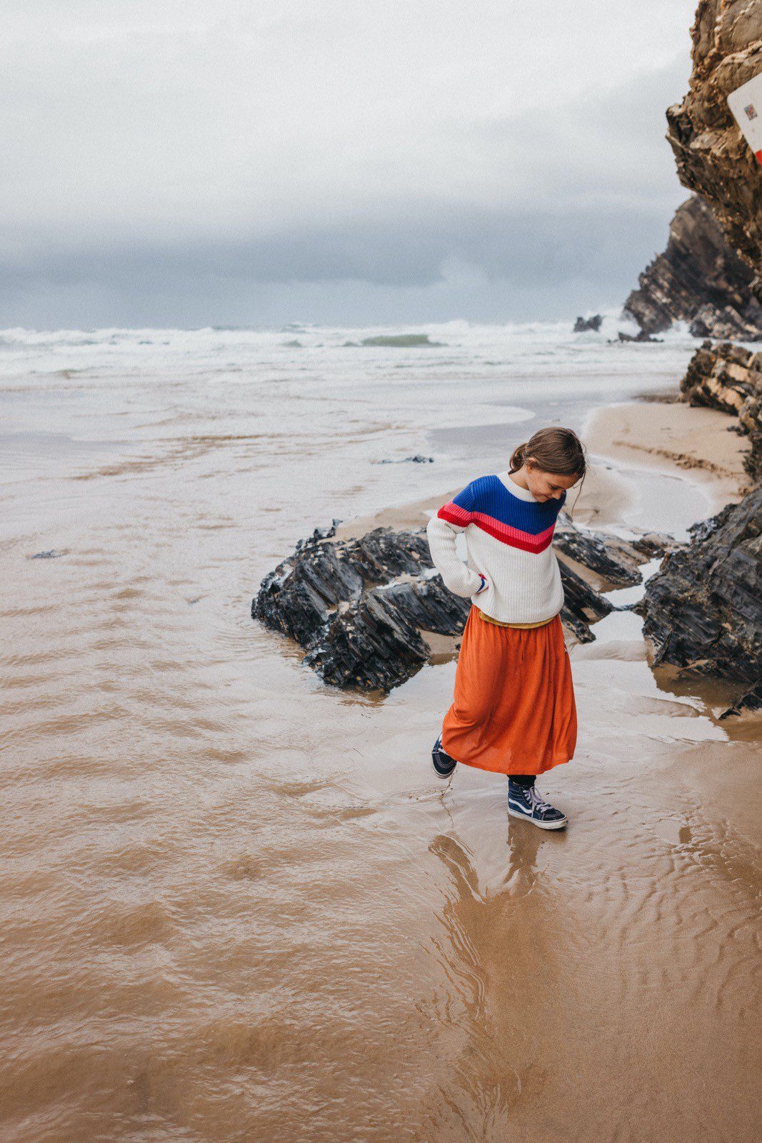 Eine Person, die einen bunten Pullover und einen orangefarbenen Rock trägt, geht an einem Sandstrand in der Nähe von dunklen Felsen entlang, während hinter ihr die Wellen schlagen - wunderschön eingefangen von Fotograf Köln unter einem dramatischen, bewölkten Himmel.