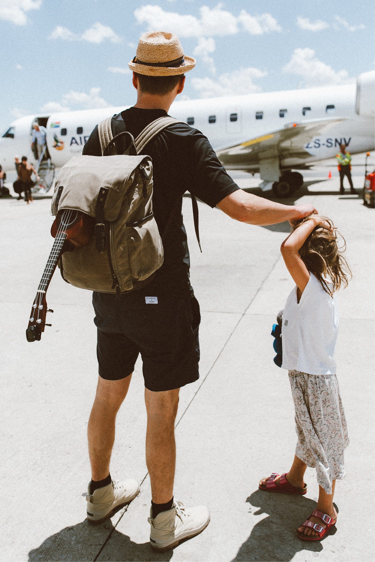 Ein Mann mit Hut und Rucksack und einer Ukulele läuft auf dem Rollfeld eines Flughafens und hält die Hand eines jungen Mädchens, während sie sich unter einem sonnigen Himmel einem kleinen Flugzeug nähern - fotografiert von Fotograf Köln mit einem Auge für herzliche Momente.