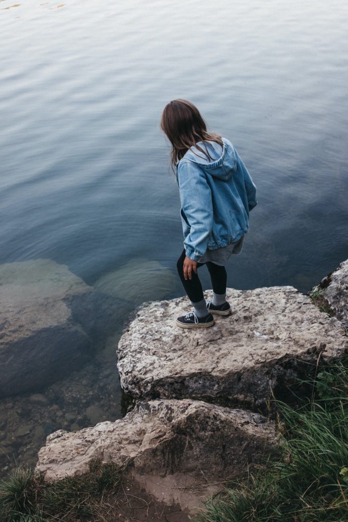 Eine Person in blauer Jacke und schwarzer Hose steht auf einem Felsvorsprung am ruhigen Wasser und blickt auf das klare Wasser und die untergetauchten Felsen hinunter - eingefangen mit dem Auge eines Werbefotografen.