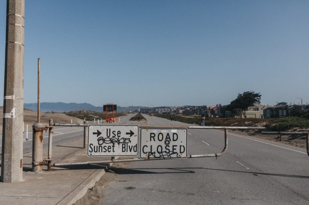 Ein "Road Closed"-Schild versperrt eine breite, leere Straße unter einem strahlend blauen Himmel. Ein anderes Schild weist die Autofahrer an, den Sunset Blvd. zu benutzen. Perfekt für einen Kids Fotograf oder Familien Fotograf, mit Gebäuden und Hügeln in der Ferne sichtbar.