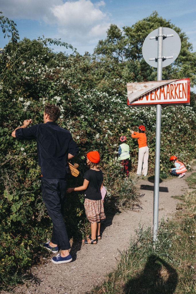 Eine Gruppe von Erwachsenen und Kindern mit roten Hüten pflückt Beeren von Sträuchern an einem sonnigen Weg. Neben ihnen steht ein Metallschild mit einem rot-weißen Schild, das teilweise von Laub verdeckt ist - perfekt für einen Kinder- oder Familienfotografen.