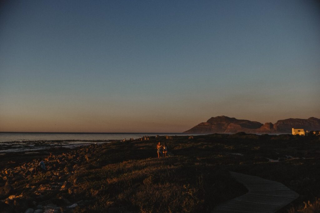 Eine Küstenlandschaft bei Sonnenuntergang mit Bergen im Hintergrund, goldenem Licht auf der Vegetation, einer hölzernen Uferpromenade und einem Paar, das in der Nähe des Ufers steht - aufgenommen von einem Werbefotografen. Der klare Himmel und die ruhige Szene strahlen Gelassenheit aus.