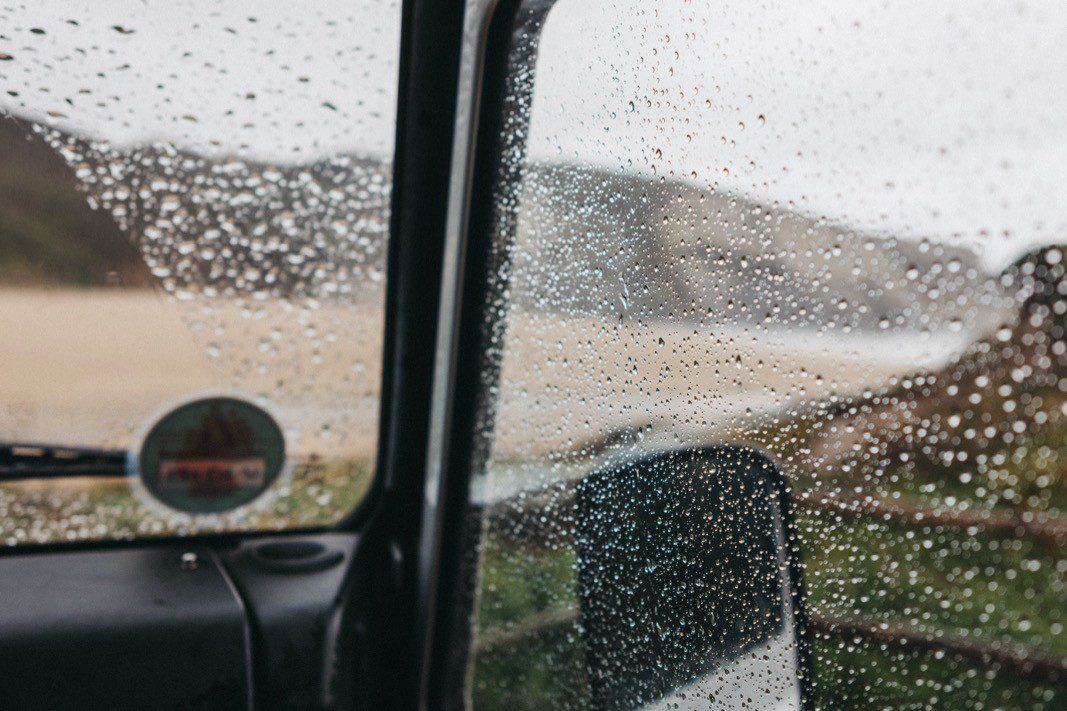 Regentropfen bedecken das Fenster eines geparkten Autos und trüben den Blick auf einen Sandstrand und die entfernten Hügel. Die von einem Fotografen Köln aufgenommene Szene sieht wolkig und ruhig aus, mit gedämpften natürlichen Farben, die durch das nasse Glas sichtbar sind.
