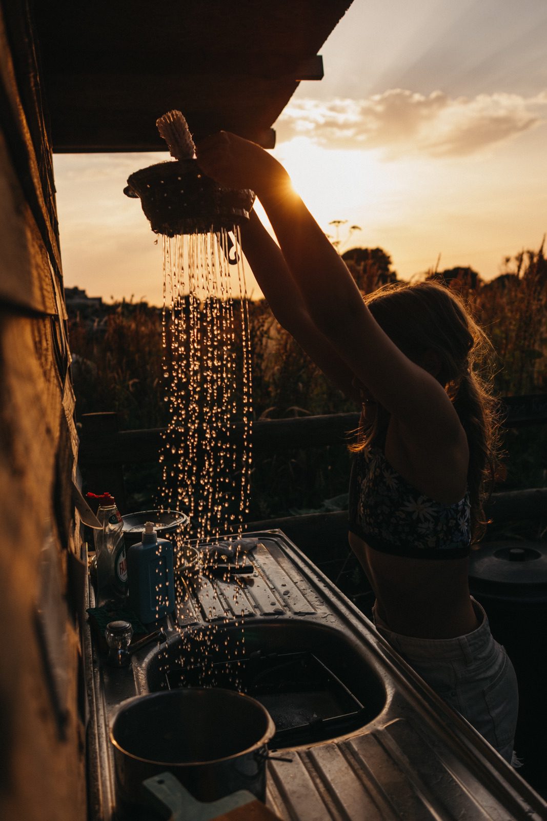 Eine Frau hält ein Sieb, durch das Wasser fließt, über eine Küchenspüle im Freien, während die Sonne untergeht. Dabei entstehen glitzernde Tropfen im goldenen Licht - eingefangen von einem Fotografen aus Köln, der ein Auge für zauberhafte Momente hat.