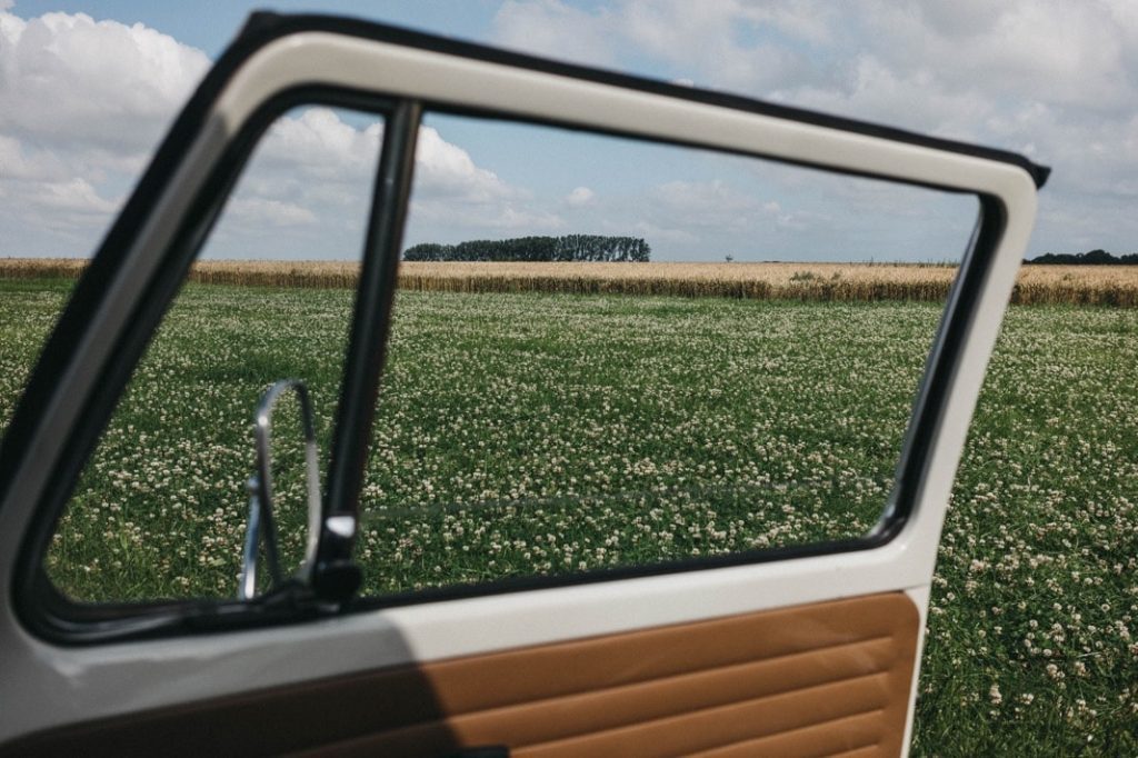 Eine offene Autotür rahmt den Blick auf ein grasbewachsenes Feld mit weißen Wildblumen ein, das sich perfekt für einen Kinderfotografen oder Werbefotografen eignet, mit Bäumen am Horizont und einem teilweise bewölkten Himmel darüber.