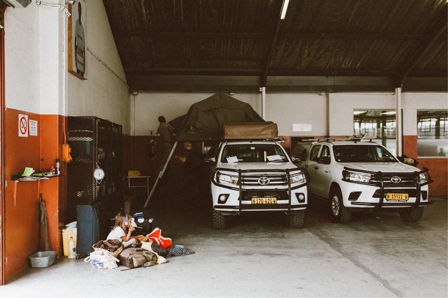 Zwei weiße Geländewagen mit Dachzelten stehen in einer Garage mit roten und weißen Wänden - perfekt für ein kreatives Fotostudio Köln oder einen Werbefotografen. Eine Person klettert auf die Leiter des Zeltes, während eine andere in der Nähe sitzt, umgeben von Taschen und Ausrüstung.