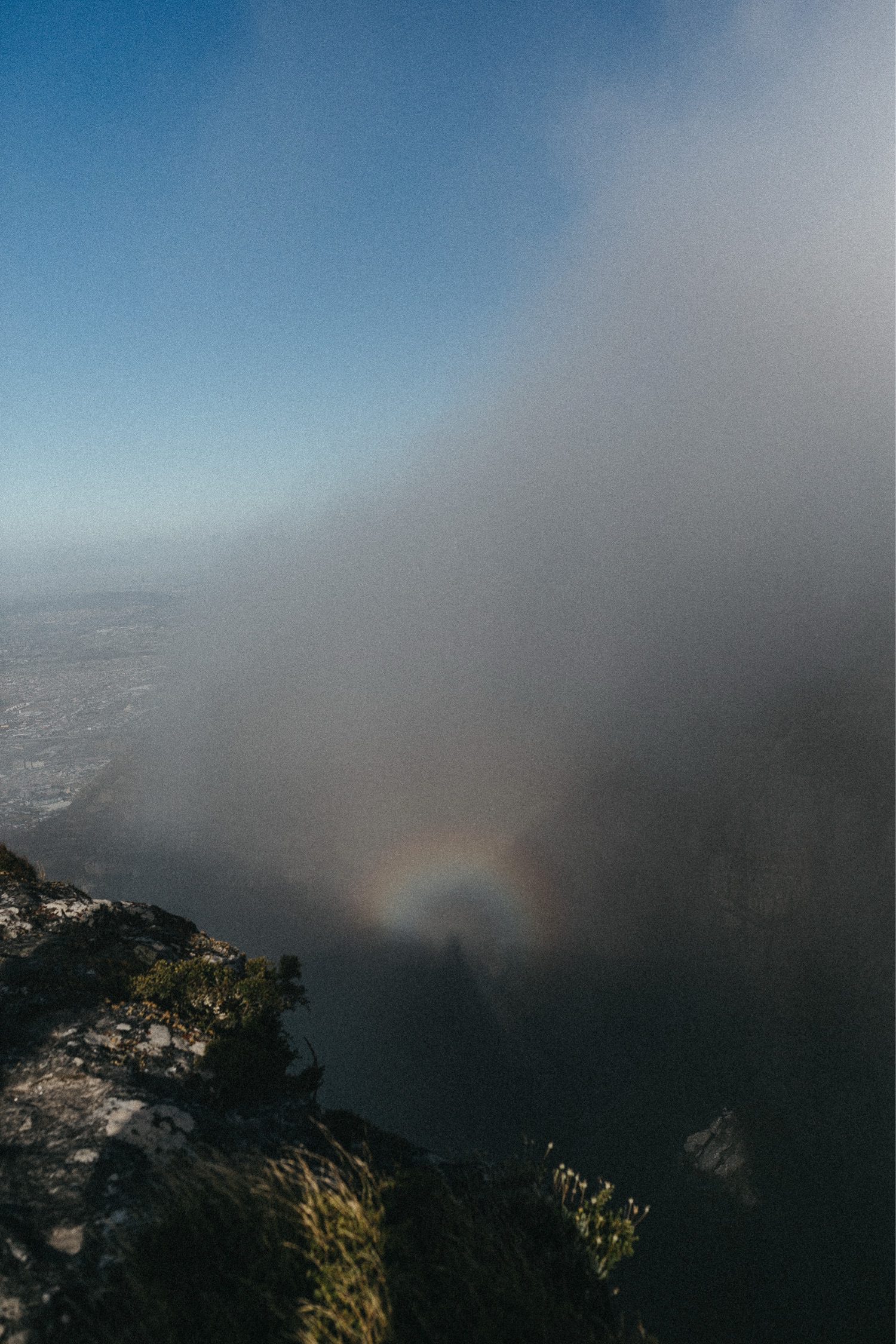 Eine neblige Berglandschaft, in der das Sonnenlicht einen schwachen, kreisförmigen Regenbogen (Glory) im Nebel erzeugt, der in seiner Mitte eine schemenhafte Figur erkennen lässt. Der Himmel ist klar und blau über den Wolken.