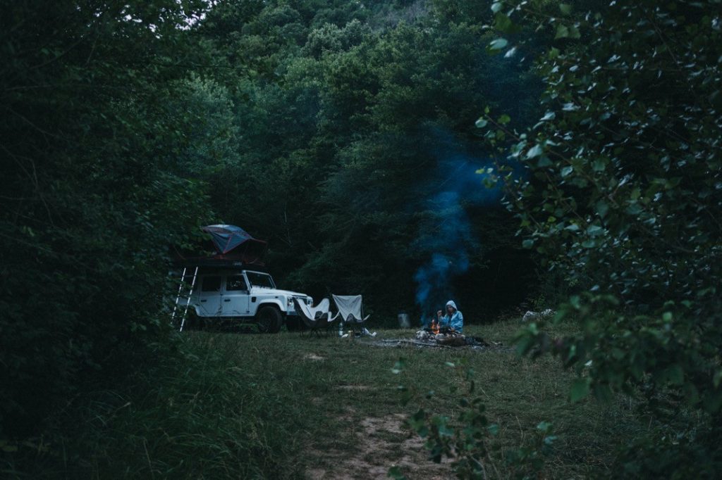 Eine Person sitzt an einem Lagerfeuer, aus dem blauer Rauch aufsteigt, in der Nähe eines weißen Geländewagens mit Dachzelt auf einer üppigen, grünen Waldlichtung. Neben dem Auto hängt Wäsche - die perfekte Inspiration für jeden Werbefotografen oder Fotografin vom Fotostudio Köln.
