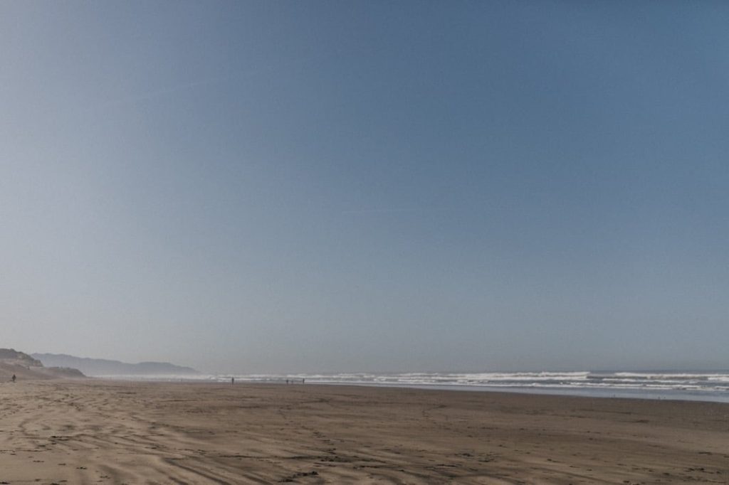 Ein breiter, leerer Sandstrand erstreckt sich in der Ferne unter einem klaren blauen Himmel, mit sanften Meereswellen auf der rechten Seite und schwachen Umrissen von Hügeln auf der linken Seite - perfekt für einen Familienfotografen, der ruhige und zeitlose Momente festhält.