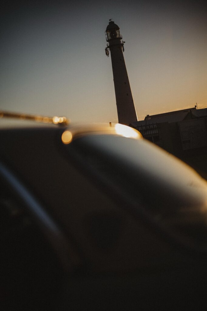 Ein hoher Leuchtturm hebt sich gegen den düsteren Himmel bei Sonnenuntergang ab, im Vordergrund sind das Dach und die Fenster eines Autos unscharf zu erkennen - aufgenommen von einem Werbefotografen des Fotostudios Köln.