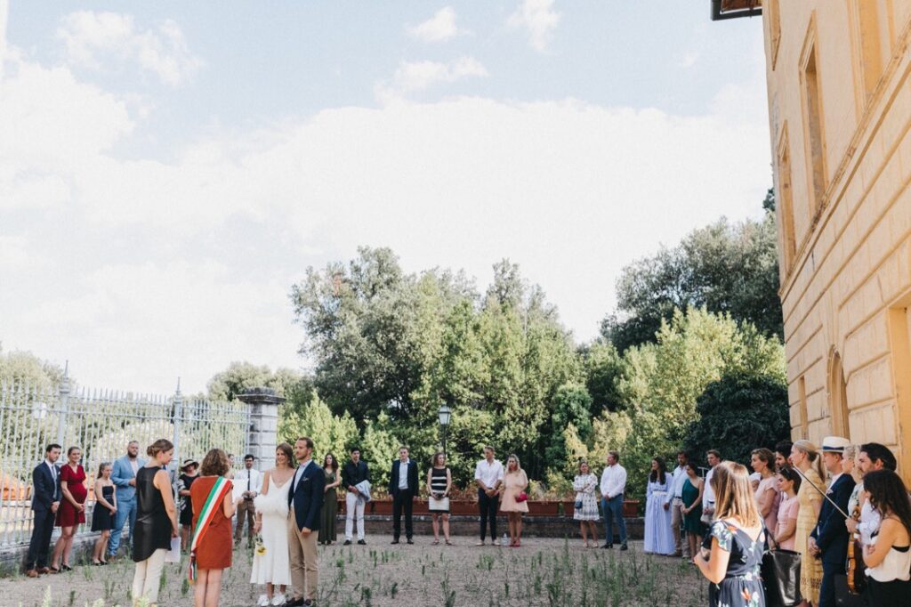 Eine Hochzeit in der Villa Cetinale findet im Freien statt, mit zwei Personen in der Mitte, umgeben von Gästen. Die Gruppe steht in der Nähe eines gelben Gebäudes, eingerahmt von üppigem Grün und einem klaren blauen Himmel im Hintergrund.
