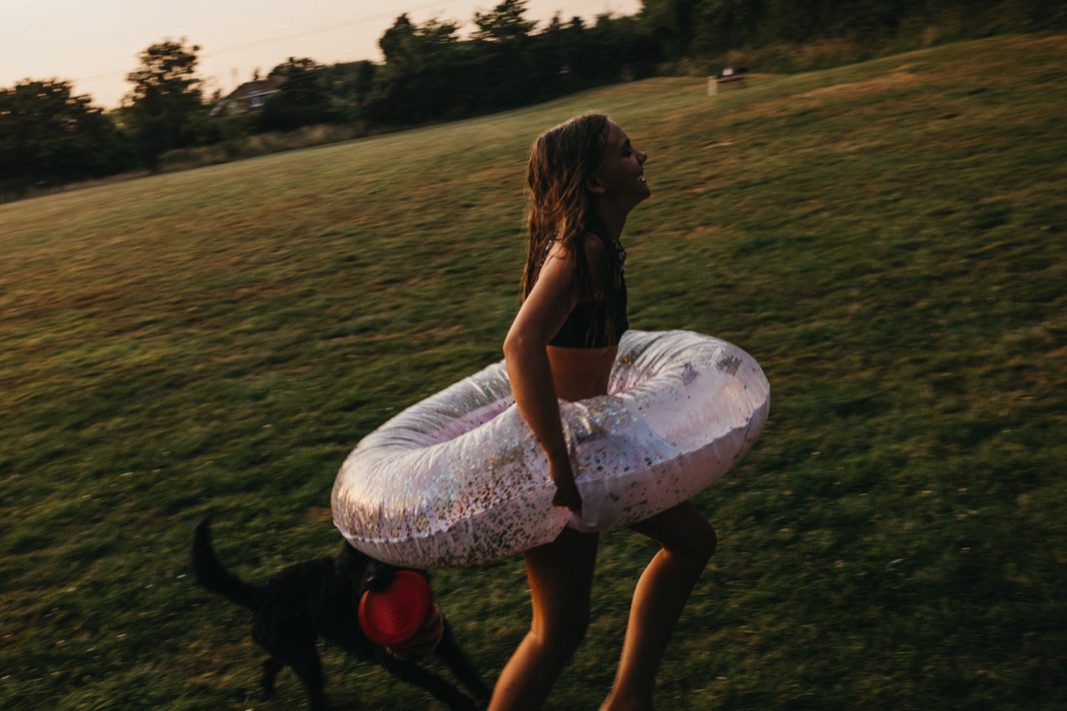 Ein Mädchen im Badeanzug und mit einem glitzernden aufblasbaren Schlauch rennt bei Sonnenuntergang über eine Wiese, gefolgt von einem schwarzen Hund mit einem roten Frisbee - fröhliche Momente mit dem kreativen Touch eines Kinderfotografen in Köln eingefangen.