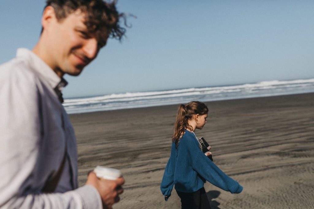 Ein Mann mit einer Tasse in der Hand und eine Frau mit einer Kamera in der Hand spazieren an einem Sandstrand entlang, unter einem klaren blauen Himmel, mit Wellen im Hintergrund - die perfekte Inspiration für jeden Familienfotografen oder Fotografin, die Momente am Meer festhält.