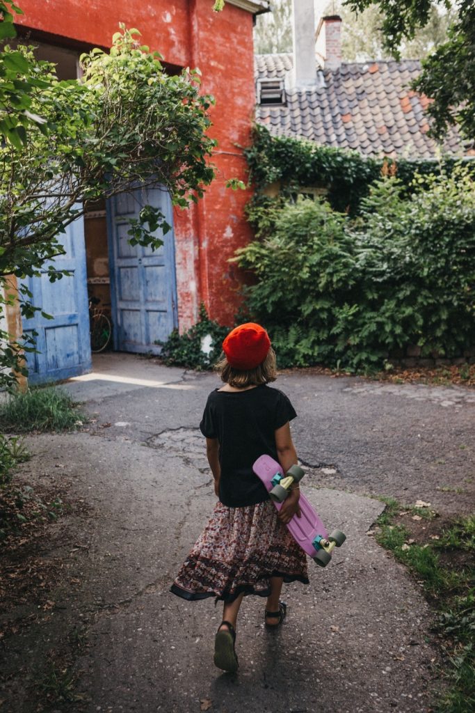 Ein Kind mit rotem Hut, schwarzem Hemd und gemustertem Rock geht auf eine offene blaue Tür zu und hält ein rosa Skateboard in einem üppigen Garten in der Hand - ein perfekter Moment, der von Kids Fotograf mit einer leuchtend roten Wand im Hintergrund eingefangen wurde.