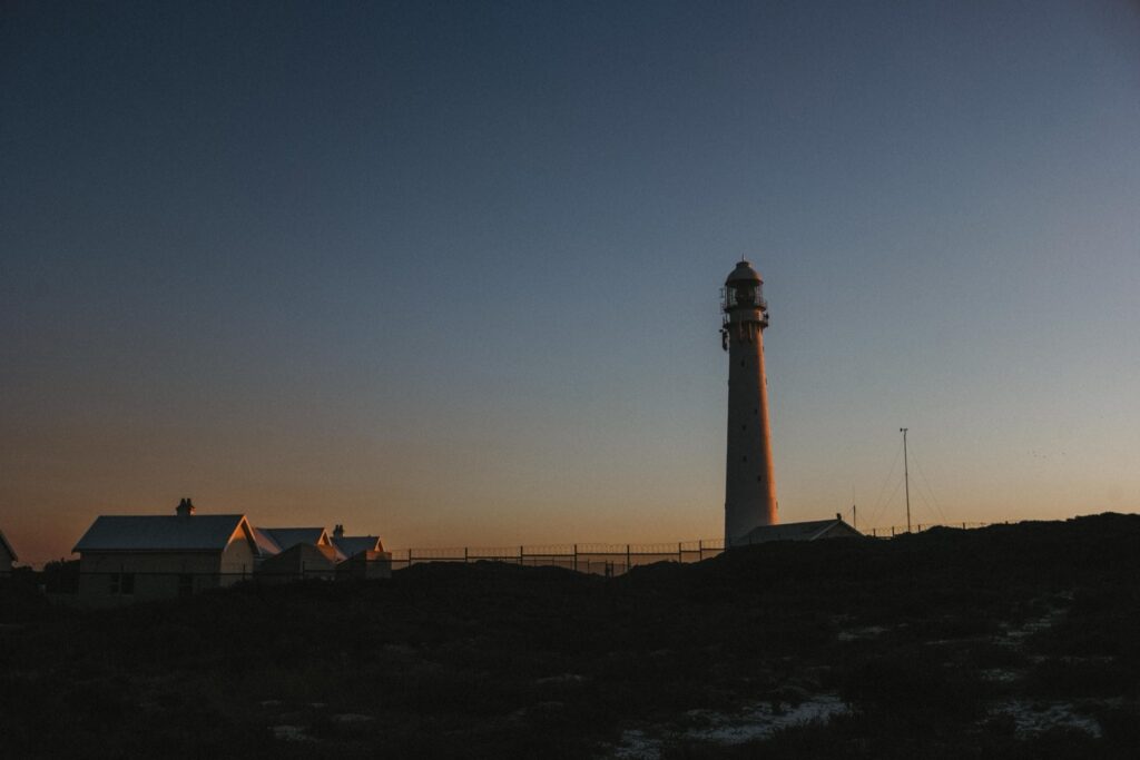 Ein hoher Leuchtturm steht bei Sonnenuntergang auf einem Hügel, in dessen Nähe sich ein paar kleine Häuser befinden. Der Himmel ist klar mit warmen orangefarbenen und blauen Tönen und wirft Silhouetten über die Landschaft - ein Motiv, das jeder Fotograf Köln lieben würde.