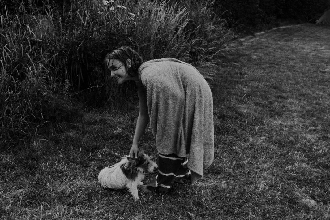 Eine in ein Handtuch gehüllte Person beugt sich hinunter, um einen kleinen, flauschigen Hund auf einem grasbewachsenen Rasen zu streicheln, mit hohen Pflanzen und Bäumen im Hintergrund. Dieses Schwarz-Weiß-Bild fängt einen zärtlichen Moment von Fotograf Köln ein.