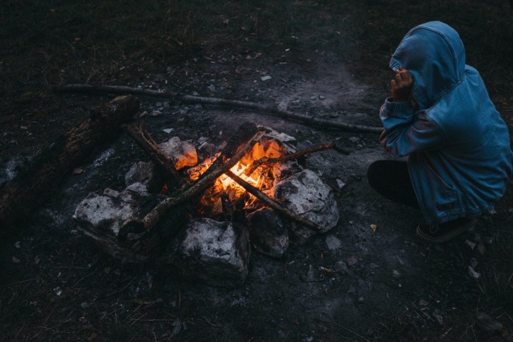 Eine Person in einem blauen Kapuzenpulli hockt neben einem kleinen Lagerfeuer, umgeben von Steinen und brennendem Holz, draußen auf einer dunklen Wiese - ein authentischer Moment, eingefangen von einer talentierten Fotografin.
