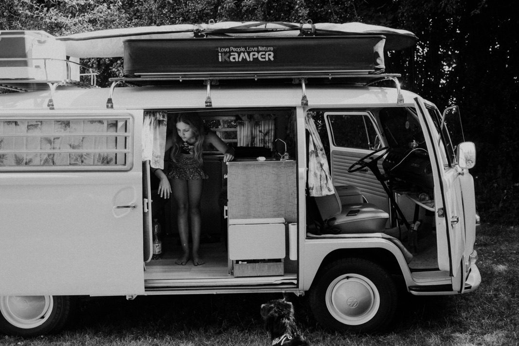 Ein Schwarz-Weiß-Foto einer talentierten Fotografin zeigt eine Frau, die in einem alten Wohnmobil mit offener Tür steht; draußen sitzt ein Hund im Gras, und im Hintergrund sind Bäume zu sehen.
