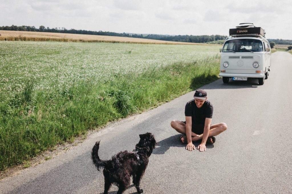 Ein Mann sitzt im Schneidersitz auf einer Landstraße und lächelt einem schwarzen Hund zu, der sich ihm nähert. In der Nähe ruht ein weißes Wohnmobil, während sich die Felder in die Ferne erstrecken - ein ruhiger Moment, den ein Familienfotograf bei teilweise bewölktem Himmel eingefangen hat.