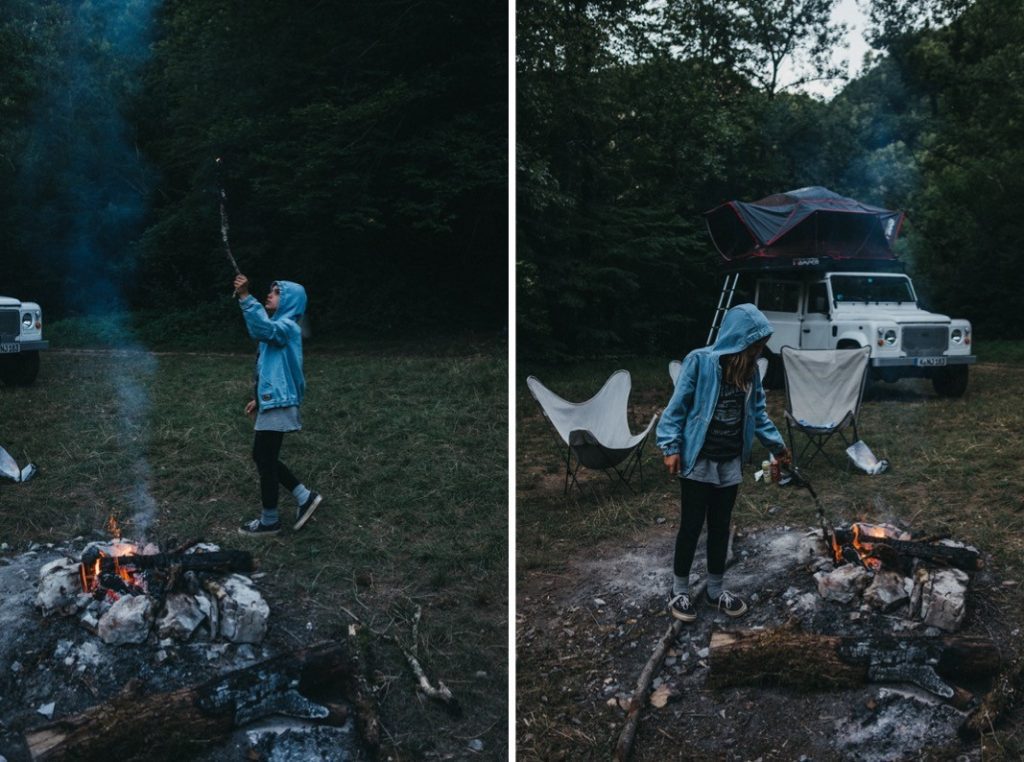 Eine Person in einer blauen Jacke steht an einem Lagerfeuer auf einem Waldcampingplatz und pflegt das Feuer mit einem Stock. Die von einem Werbefotografen aufgenommene Szene zeigt einen weißen Geländewagen mit Dachzelt und zwei Campingstühle, die in der Nähe aufgestellt sind.