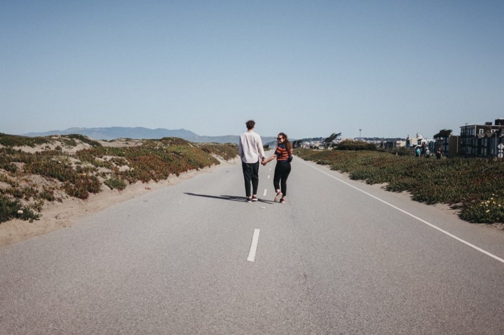 Ein Paar spaziert Hand in Hand mitten auf einer leeren Straße, umgeben von Sanddünen und niedrigem Grün unter einem klaren blauen Himmel - wunderschön eingefangen von einem Familienfotografen aus Köln.