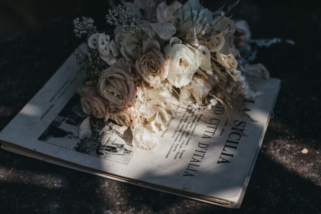 Ein Strauß getrockneter Blumen ruht auf einem alten, leicht vergilbten Buch oder einem Blatt Papier mit italienischem Text - perfekt für eine sizilianische Hochzeit -, das in sanftes, natürliches Licht getaucht ist und eine kleine Schwarz-Weiß-Illustration zeigt.