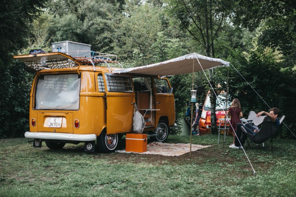 Ein gelber Oldtimer-Wohnwagen mit Vorzelt ist auf einer Wiese geparkt. Zwei Personen entspannen sich auf Stühlen unter Bäumen, während die Campingausrüstung neben dem Van aufgebaut ist - die perfekte Inspiration für einen Familienfotografen inmitten von üppigem Grün.