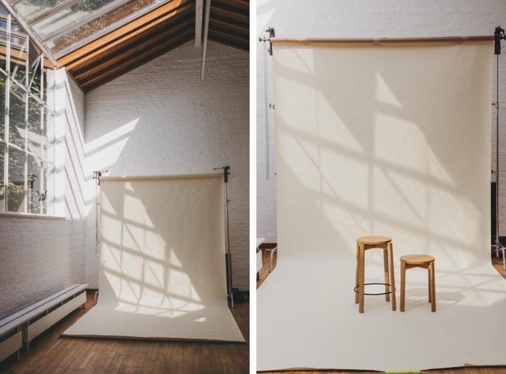 In einem hellen Studio mit Holzfußboden und weißen Backsteinwänden ist ein farbneutraler Hintergrund aufgebaut - ideal für einen Familienfotografen. Zwei Holzhocker, ein hoher und ein kurzer, stehen vor der Kulisse, während das Sonnenlicht gitterartige Schatten wirft.